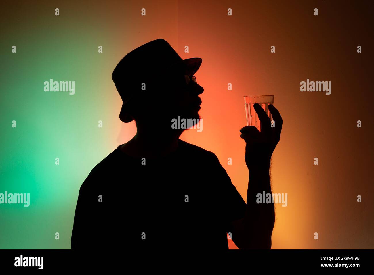 Uomo con cappello in silhouette che regge un bicchiere d'acqua su sfondo colorato. Foto Stock