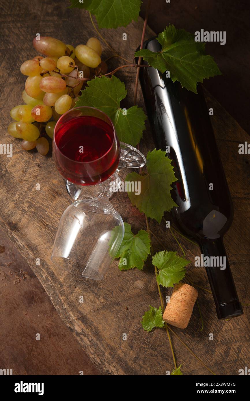 Il concetto di una deliziosa bevanda alcolica, vino e uva. Bottiglia di vino, mazzo di uva, bicchiere vuoto e bicchiere di vino rosso su legno rustico scuro Foto Stock