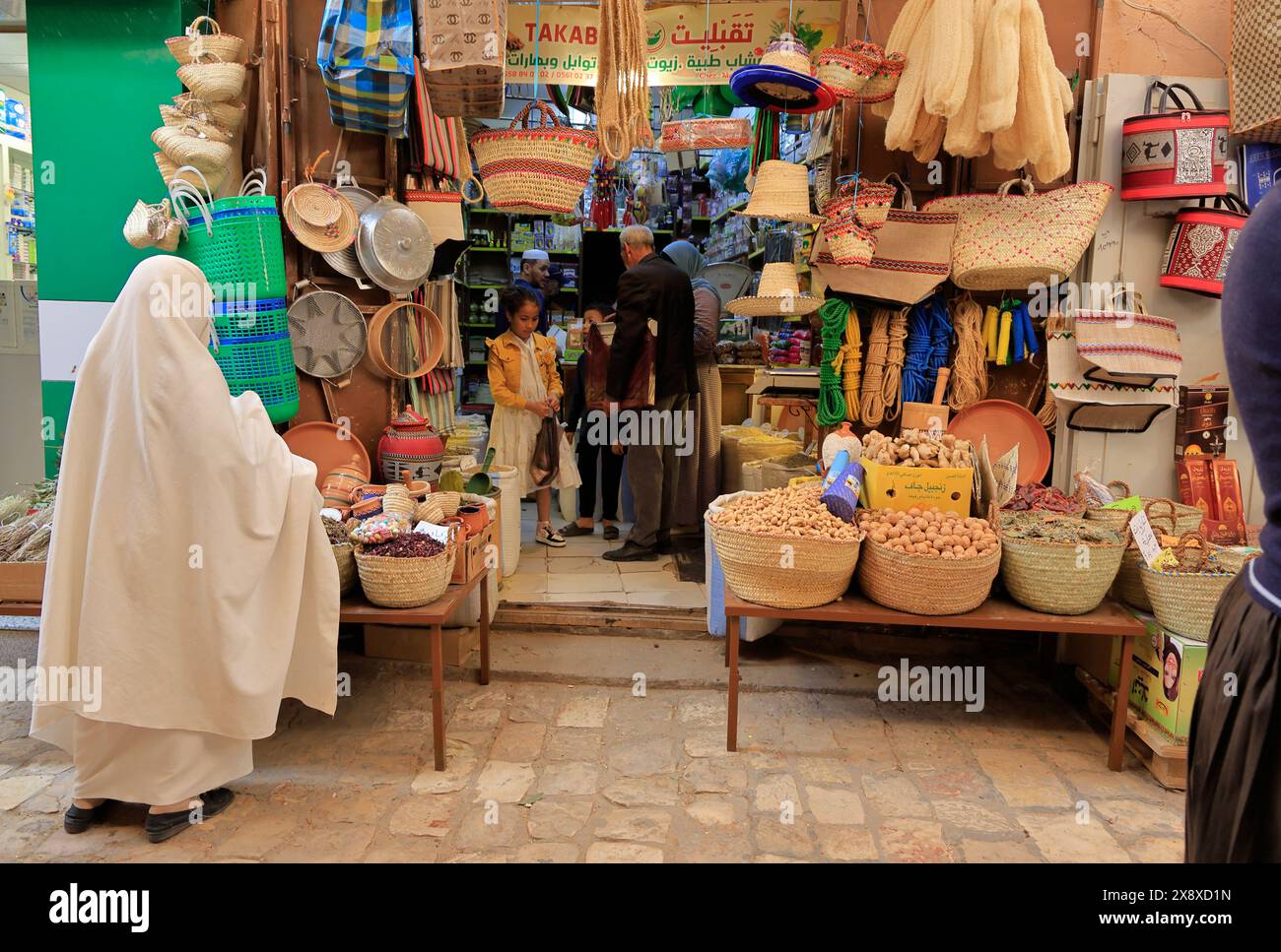 Una donna velata a haik bianca che fa shopping di fronte a un negozio di articoli per la casa e alimentari nel mercato di Ghardaia. Valle di Mzab. Provincia di Ghardaia. Sahara settentrionale. Algeria Foto Stock