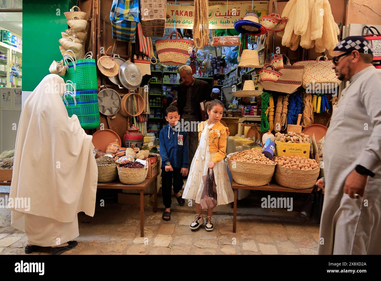 Gente Mozabite e bambini che fanno shopping nel mercato di Ghardaia con un negozio di articoli per la casa e alimentari sullo sfondo.Ghardaia.Valle di Mzab. Provincia di Ghardaia. Sahara settentrionale. Algeria Foto Stock