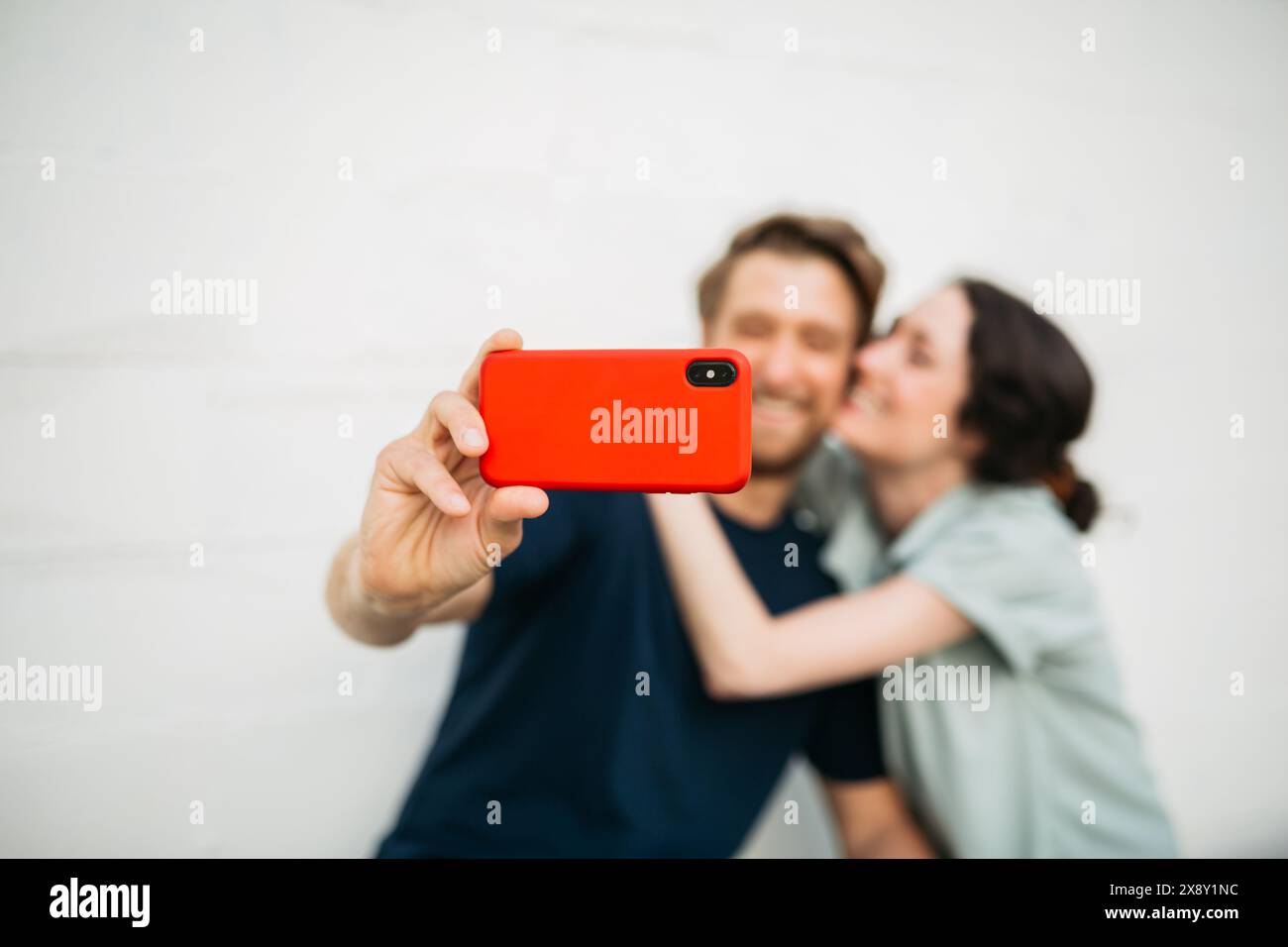 Coppia seduta insieme, sorridente e scattata un selfie con un telefono rosso contro un muro bianco. Le loro espressioni mostrano felicità e vicinanza. Foto Stock