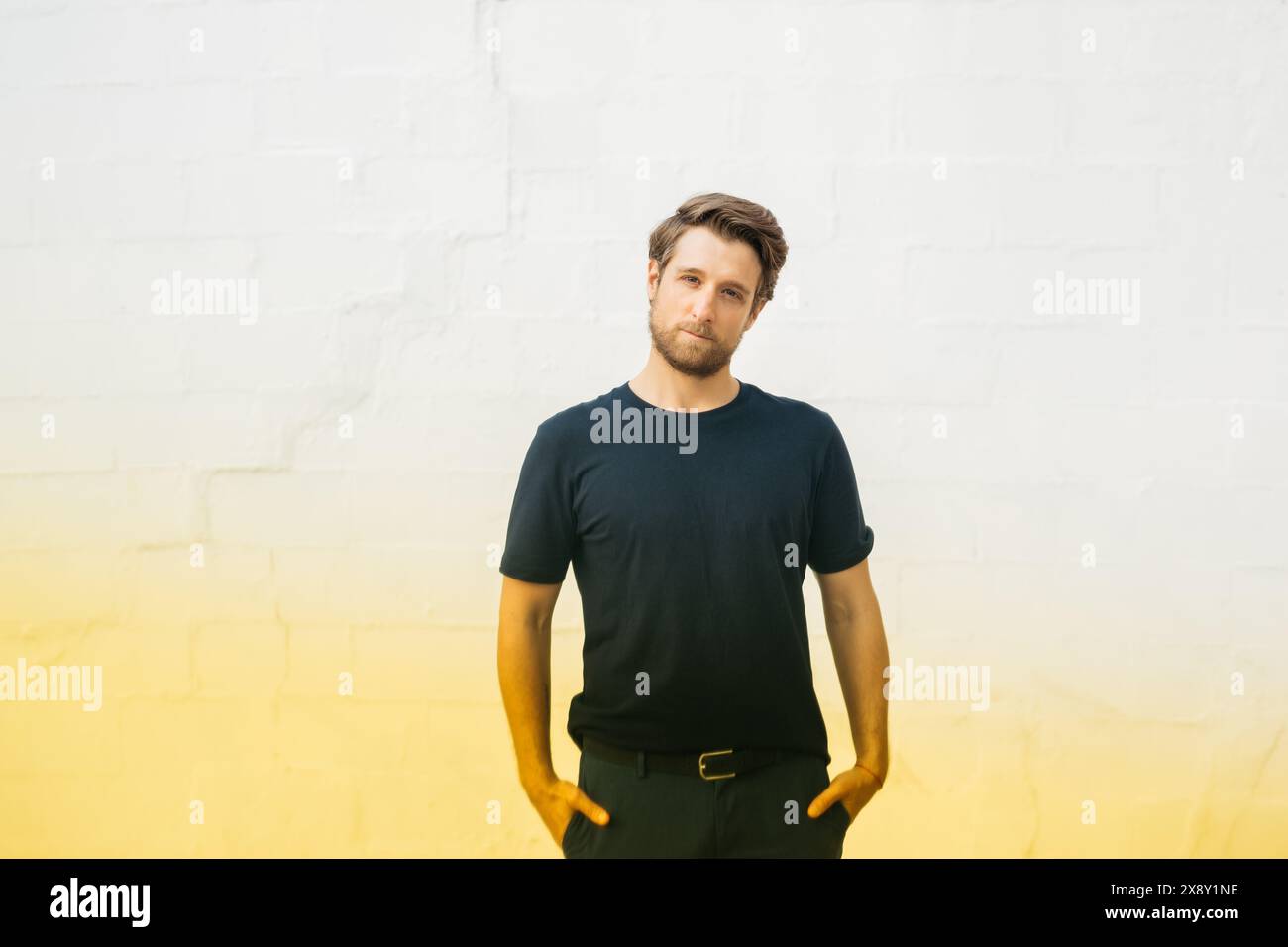 Ritratto di un uomo in piedi con le mani in tasca, con una camicia scura e un jeans, su uno sfondo bianco in mattoni. Foto Stock