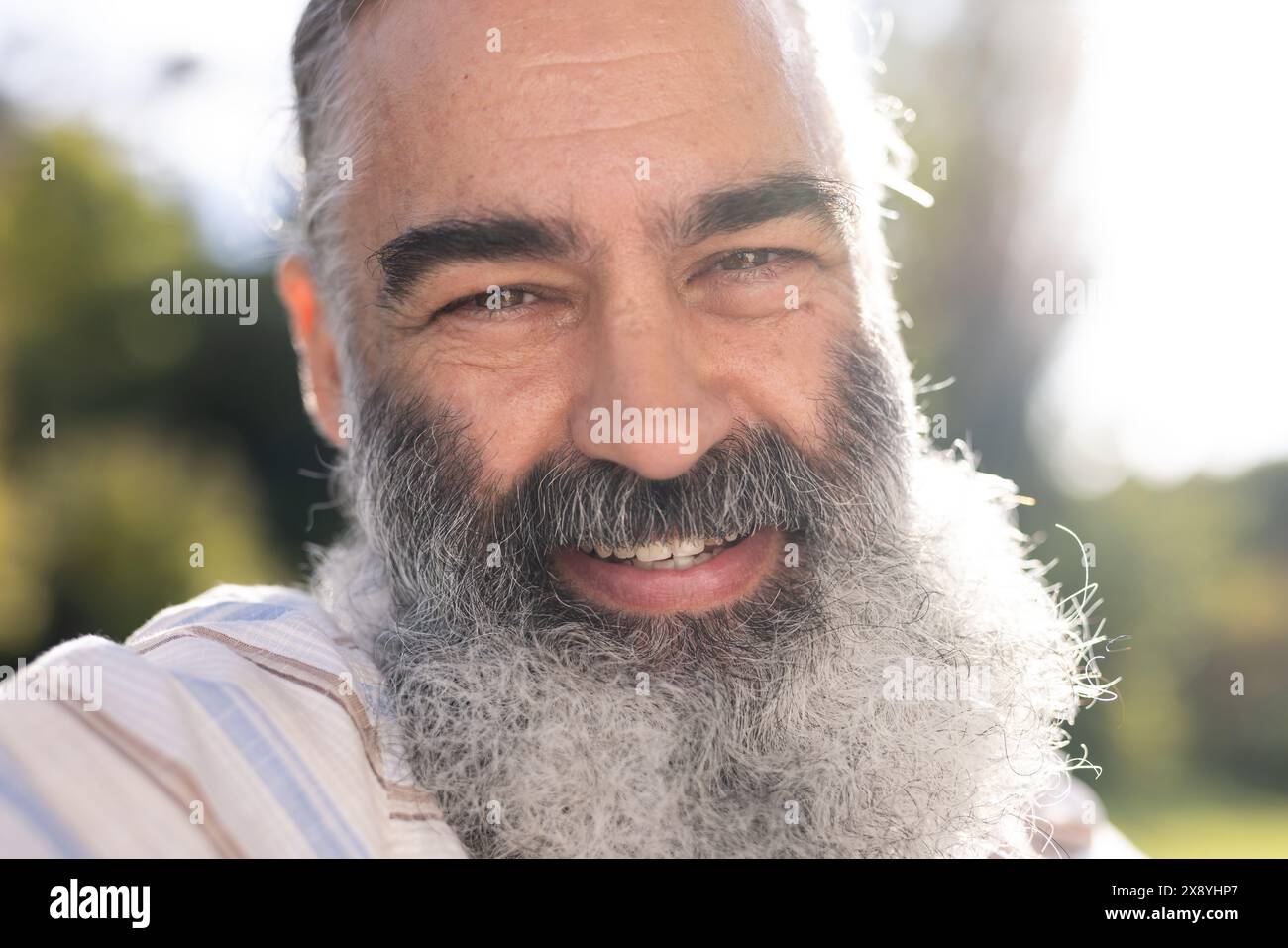 Uomo anziano caucasico con barba grigia, indossa una camicia a righe chiare e sorride all'aperto Foto Stock