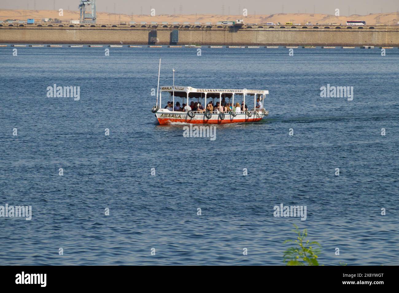 Barca turistica con la diga bassa di Assuan alle spalle. Assuan, Egitto Foto Stock