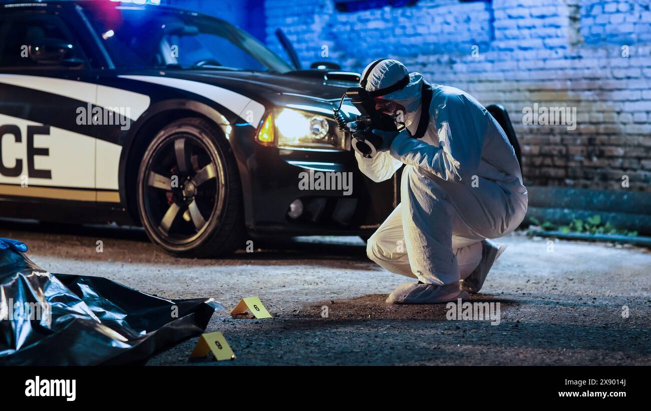 Specialista forense che scatta foto del cadavere in una borsa per il corpo su una scena del crimine. Foto serale di un ufficiale della squadra di polizia professionista in tuta da tuta con fotocamera che documenta indizi e tracce Foto Stock