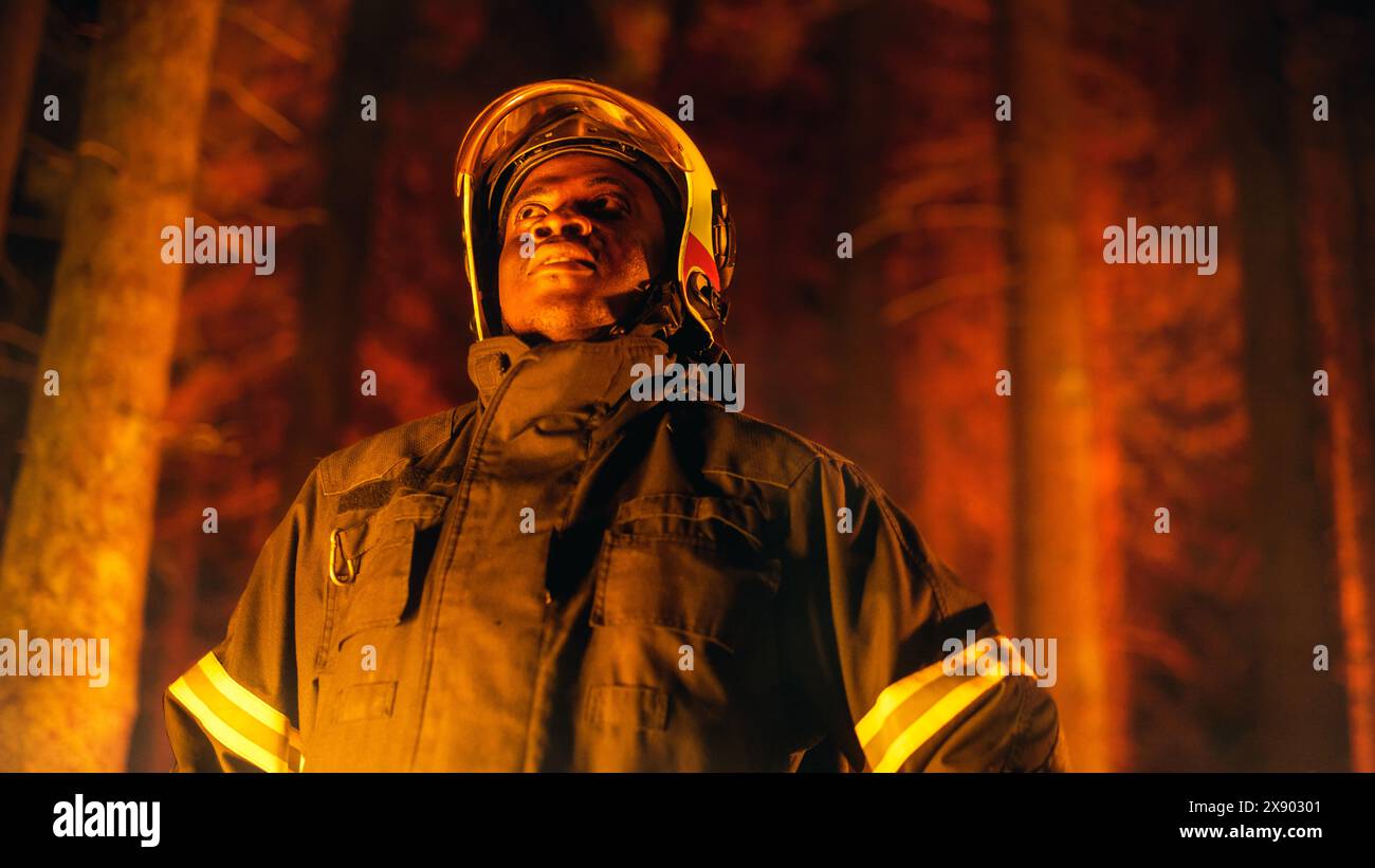 Coraggioso giovane combattente afroamericano dei vigili del fuoco con l'uniforme di sicurezza e un casco che guarda intorno alla foresta durante un incendio. Leader di squadra professionista che valuta la situazione pericolosa. Foto Stock