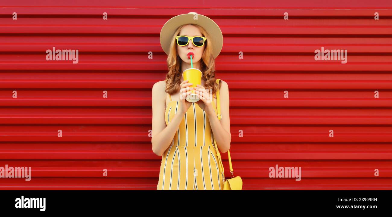 Una giovane donna bella ed elegante che beve un succo fresco che regge una tazza con cappello estivo, un vestito giallo che posa sullo sfondo rosso della parete Foto Stock