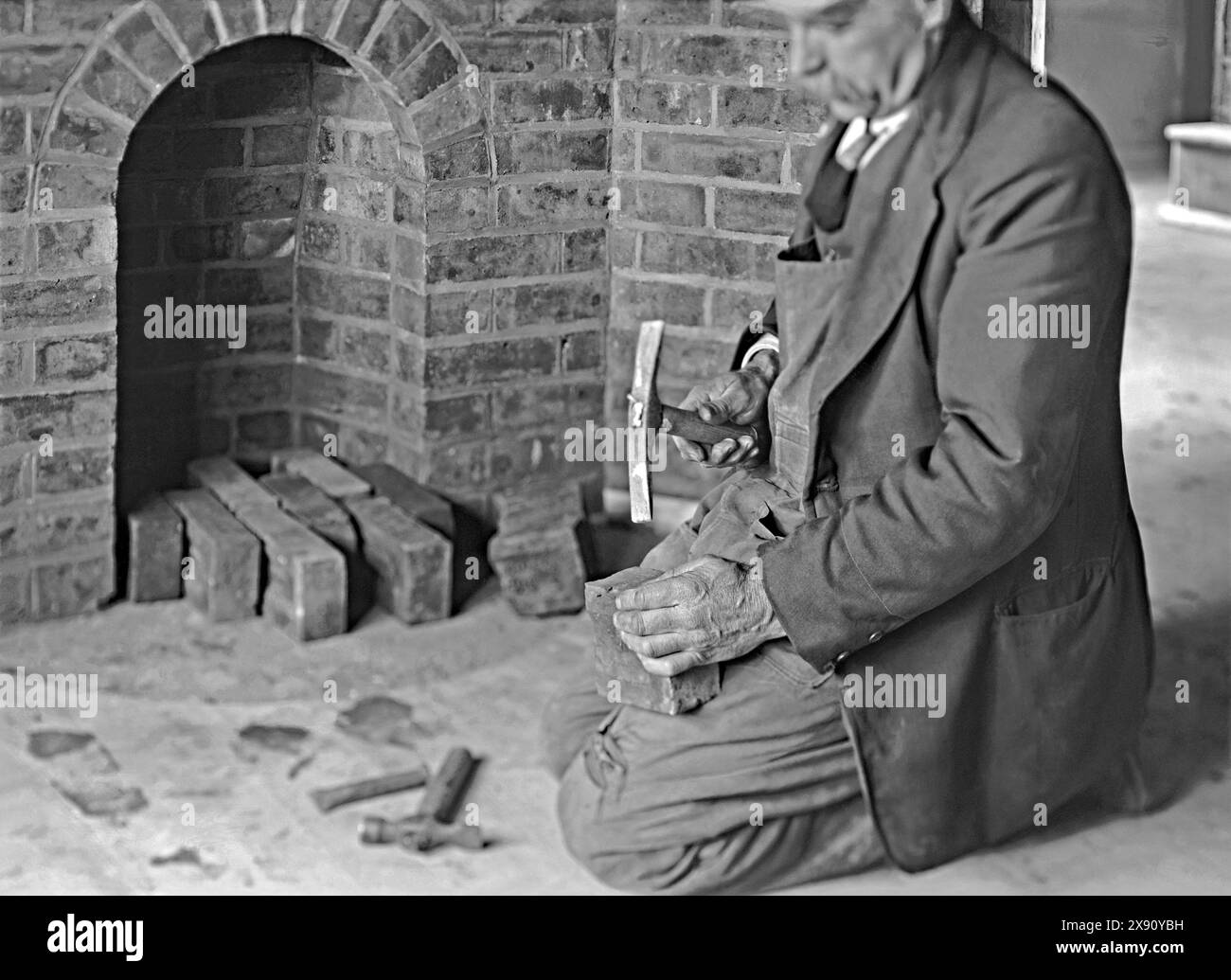 Un operaio edile che taglia e forma un mattone utilizzando un martello per mattoni o un martello di «scutch». I lavori sono stati svolti in una casa a Kennel Lane, Fetcham, Leatherhead, Surrey, Inghilterra, Regno Unito c. 1930. I martelli per mattoni sono utilizzati per dividere i mattoni in modo pulito e tritare («Unione») qualsiasi mattone in eccesso utilizzando la «lama» dello scalpello della testa. Qui un martello e uno scalpello da muratura, utilizzati per effettuare un taglio iniziale su un mattone, si trovano sul pavimento - una fotografia vintage anni '1920/'30. Foto Stock