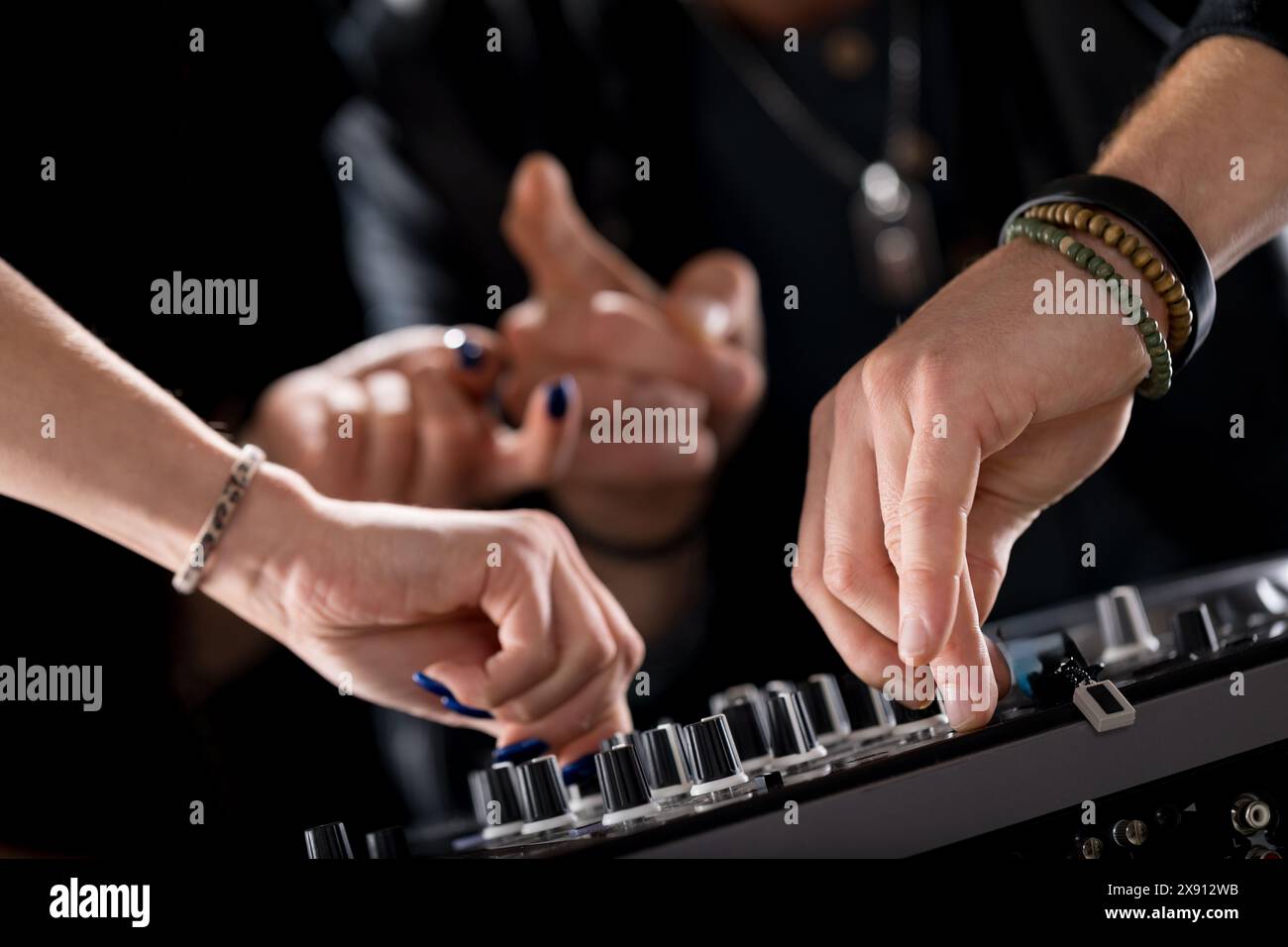 Primo piano di mani che lavorano su un mixer DJ, regolano le manopole, mentre altre mani gestiscono lo sfondo. La scena enfatizza precisione, controllo, Foto Stock