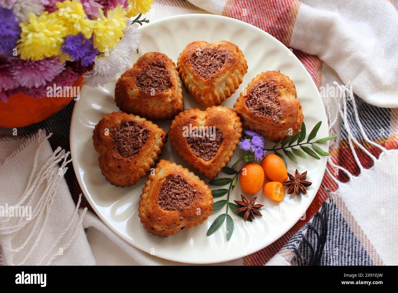 Biscotti a forma di cuore con scaglie di cioccolato disposti su un piatto con fiori, kumquat e anice stellato, su un panno a quadri Foto Stock