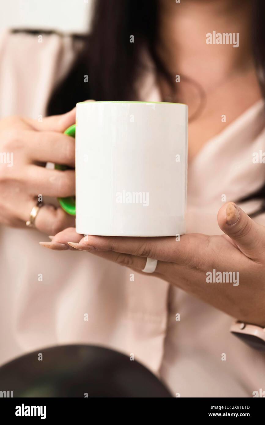 Donna che tiene in mano una tazza bianca con manico verde, anelli e camicetta di colore chiaro Foto Stock