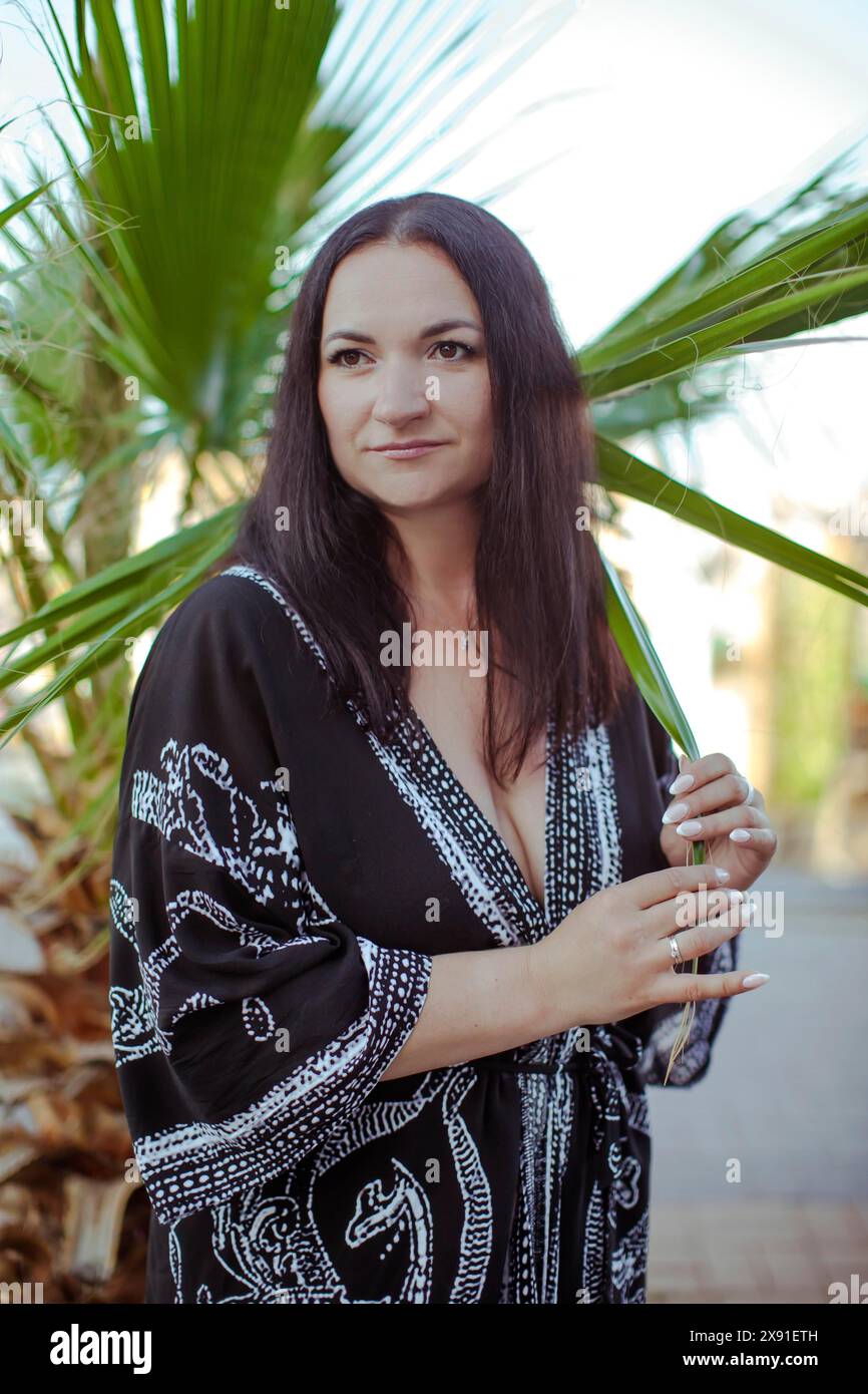 Una donna con i capelli scuri in un abito nero si trova all'aperto vicino a una palma, sembra rilassata e sorridente, Turchia, Antalya Foto Stock