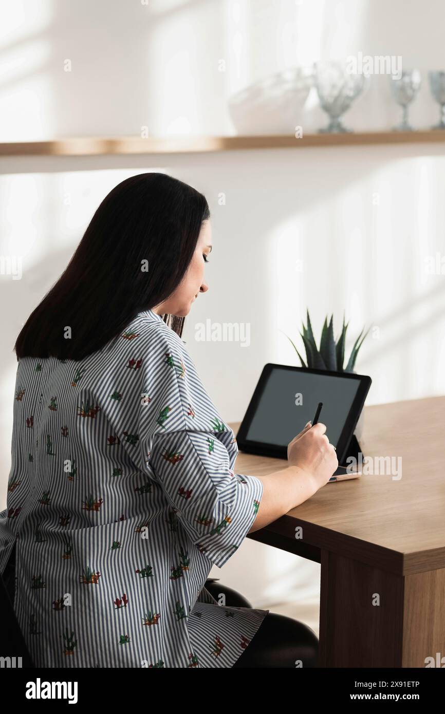 Una donna con una camicia a righe utilizza uno stilo su un tablet su un tavolo di legno con luci e ombre in un interno minimalista Foto Stock
