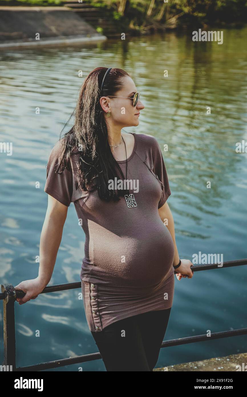 Una donna incinta che indossa occhiali da sole e un abbigliamento casual si trova accanto a una ringhiera vicino all'acqua, riflettendo un'atmosfera tranquilla. Bielorussia, Minsk Foto Stock