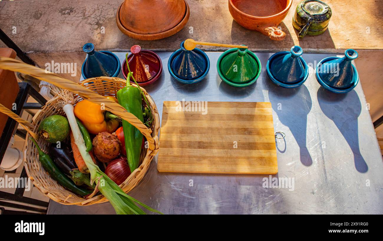 Un vivace cestino di verdure fresche accanto a colorate pentole di tagine su un tagliere di legno in un ambiente rustico da cucina all'aperto. Foto Stock