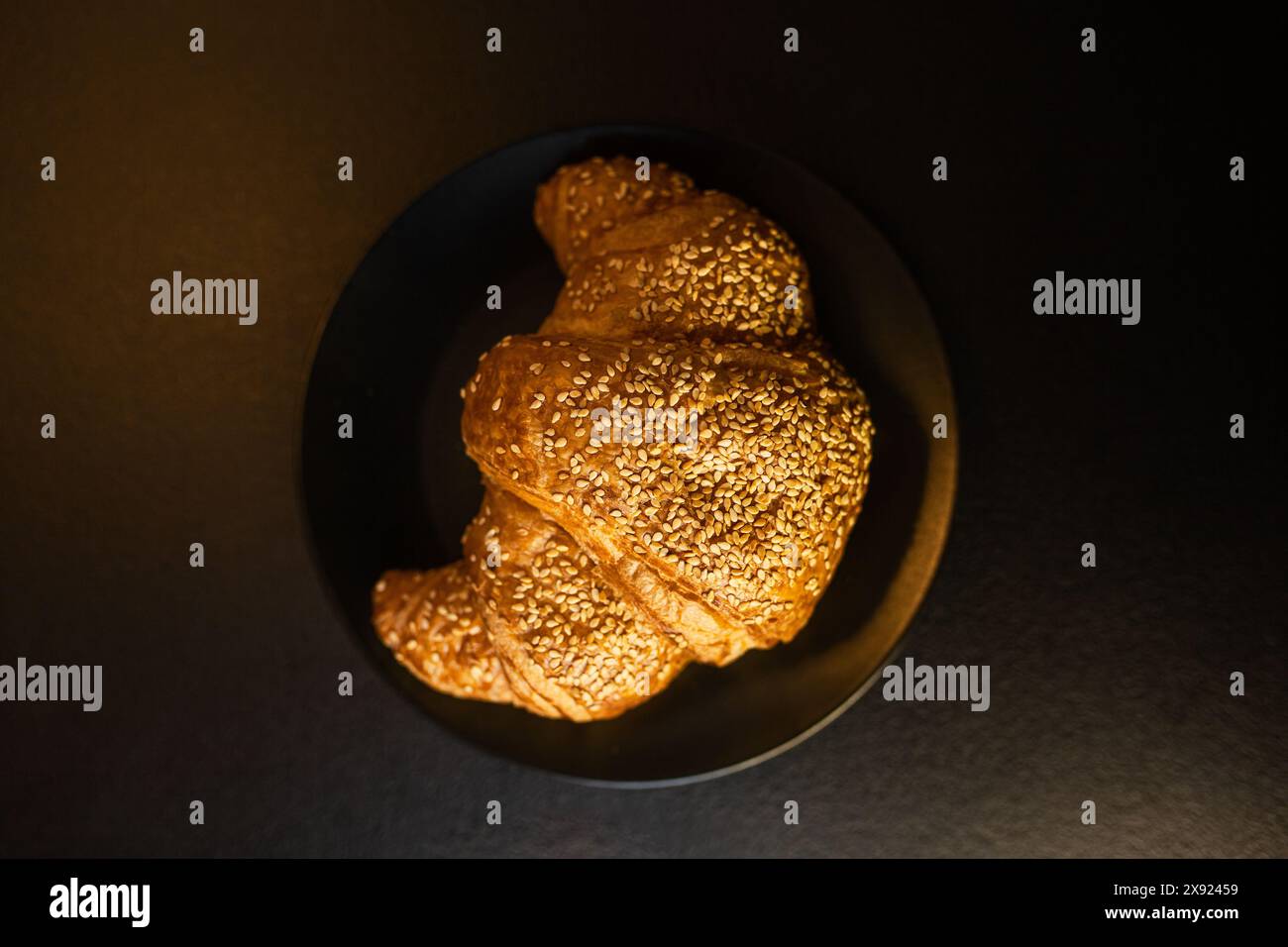 Un croissant di sesamo appena sfornato servito su un piatto scuro sotto un'illuminazione soffusa, evidenziando deliziose consistenze da forno. Foto Stock