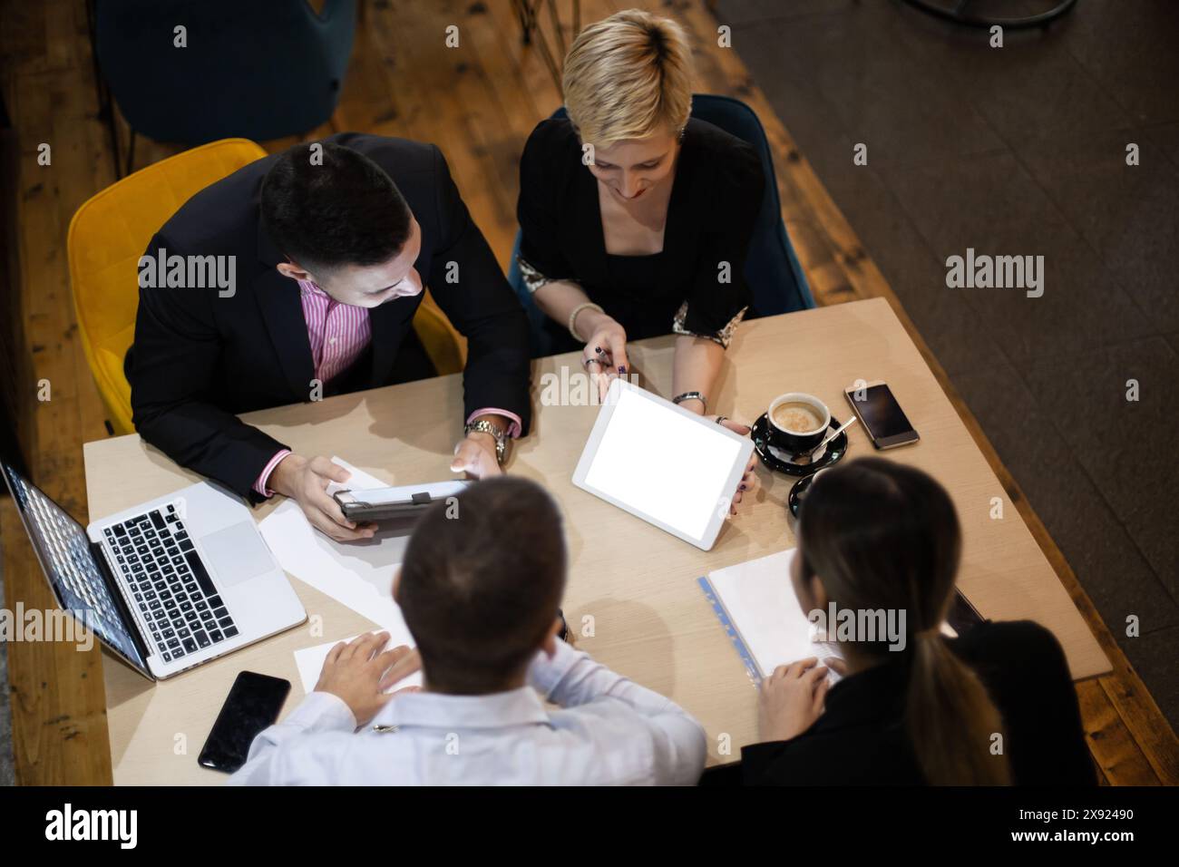 Quattro professionisti aziendali discutono la strategia a una tavola rotonda con i dispositivi digitali e il caffè in un ambiente di lavoro elegante. Foto Stock