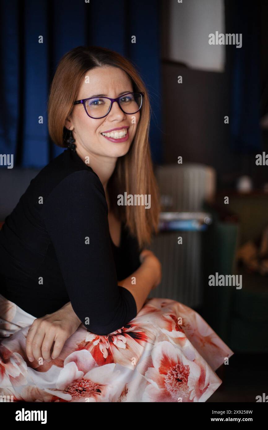 Ritratto di una donna gioiosa che indossa gli occhiali, sorride alla fotocamera mentre è seduta in una stanza calda e poco illuminata, che trasmette un senso di comfort e felicità. Foto Stock