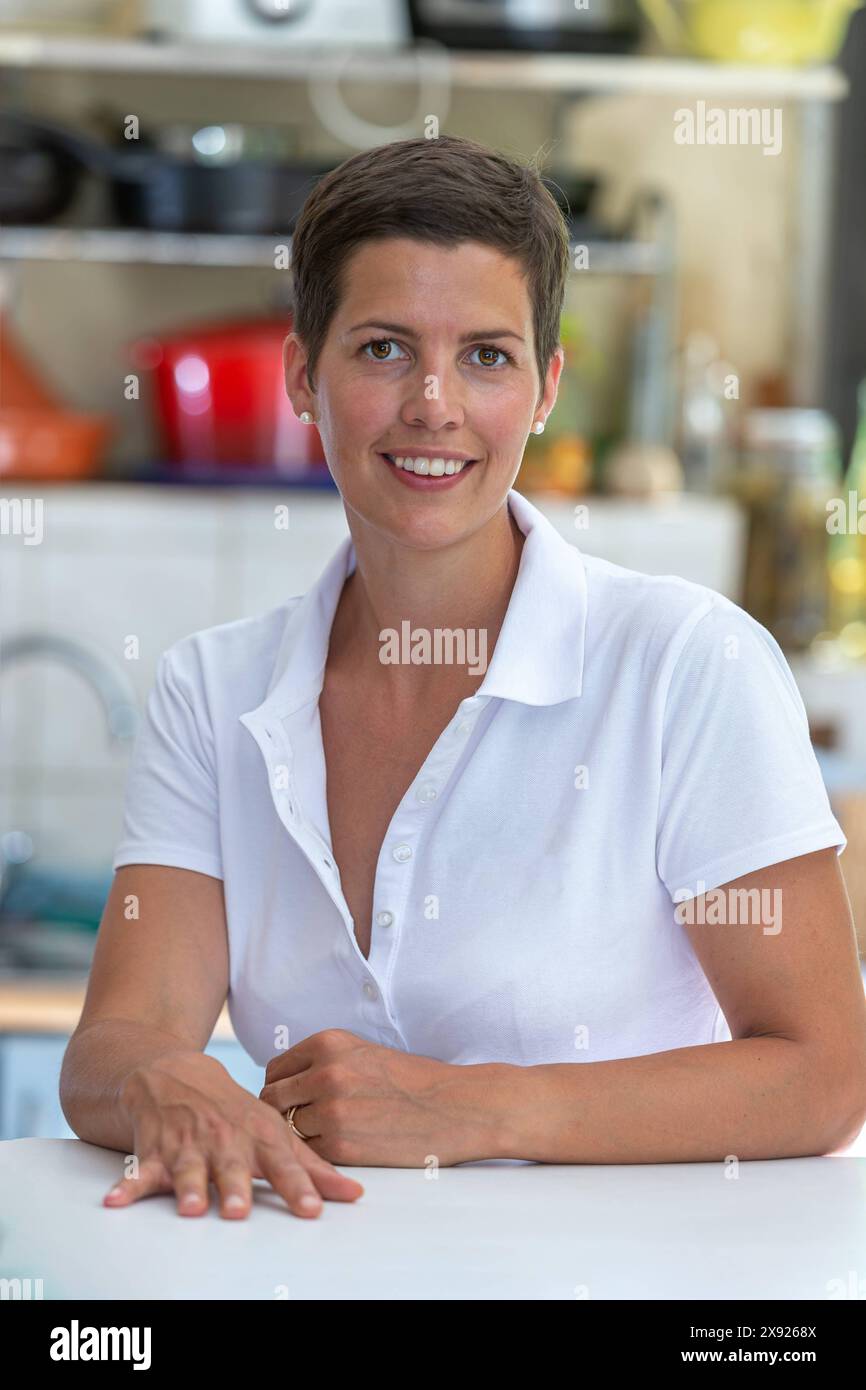 Giovane donna bruna con capelli corti appoggiati su un tavolo, utensili da cucina sullo sfondo. Ritratto di una giovane donna in cucina 016643 092 Foto Stock