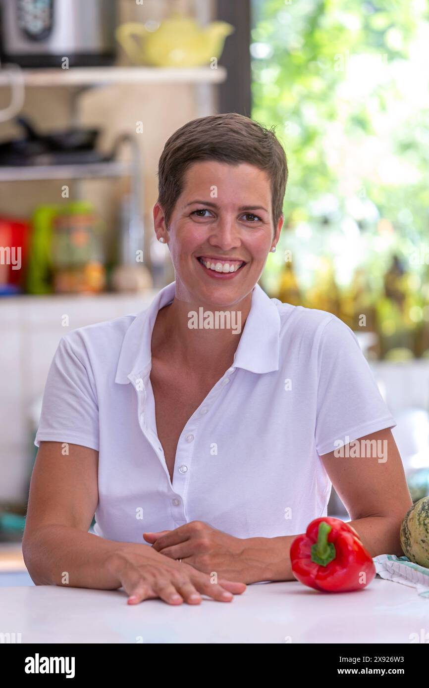 Giovane donna bruna con capelli corti appoggiati su un tavolo, utensili da cucina sullo sfondo. Ritratto di una giovane donna in cucina 016643 093 Foto Stock