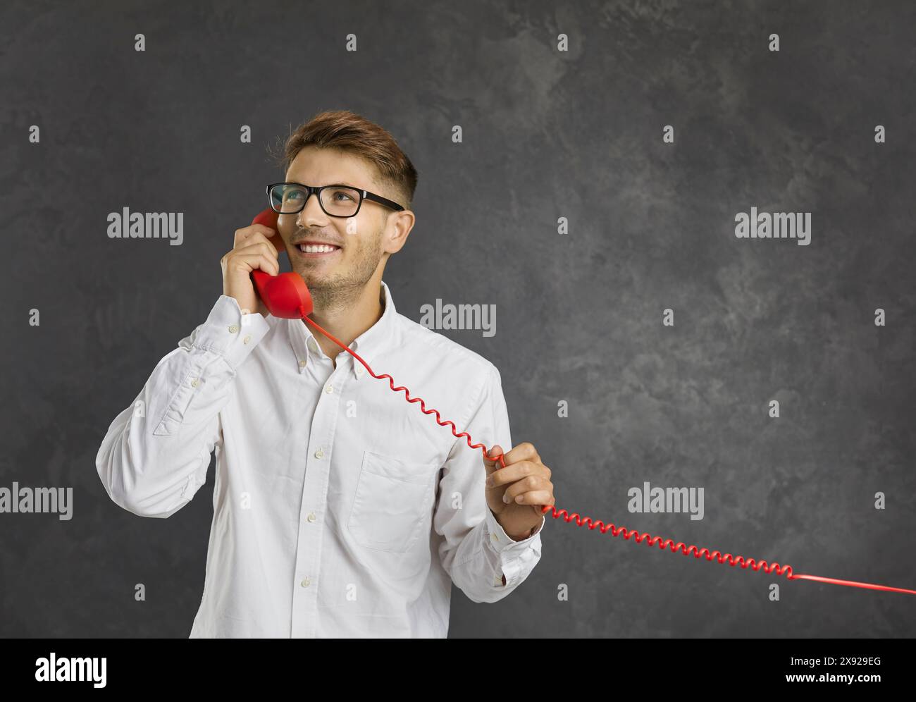 Ritratto di un giovane uomo felice e sognante che parla su un telefono fisso su uno sfondo grigio. Foto Stock