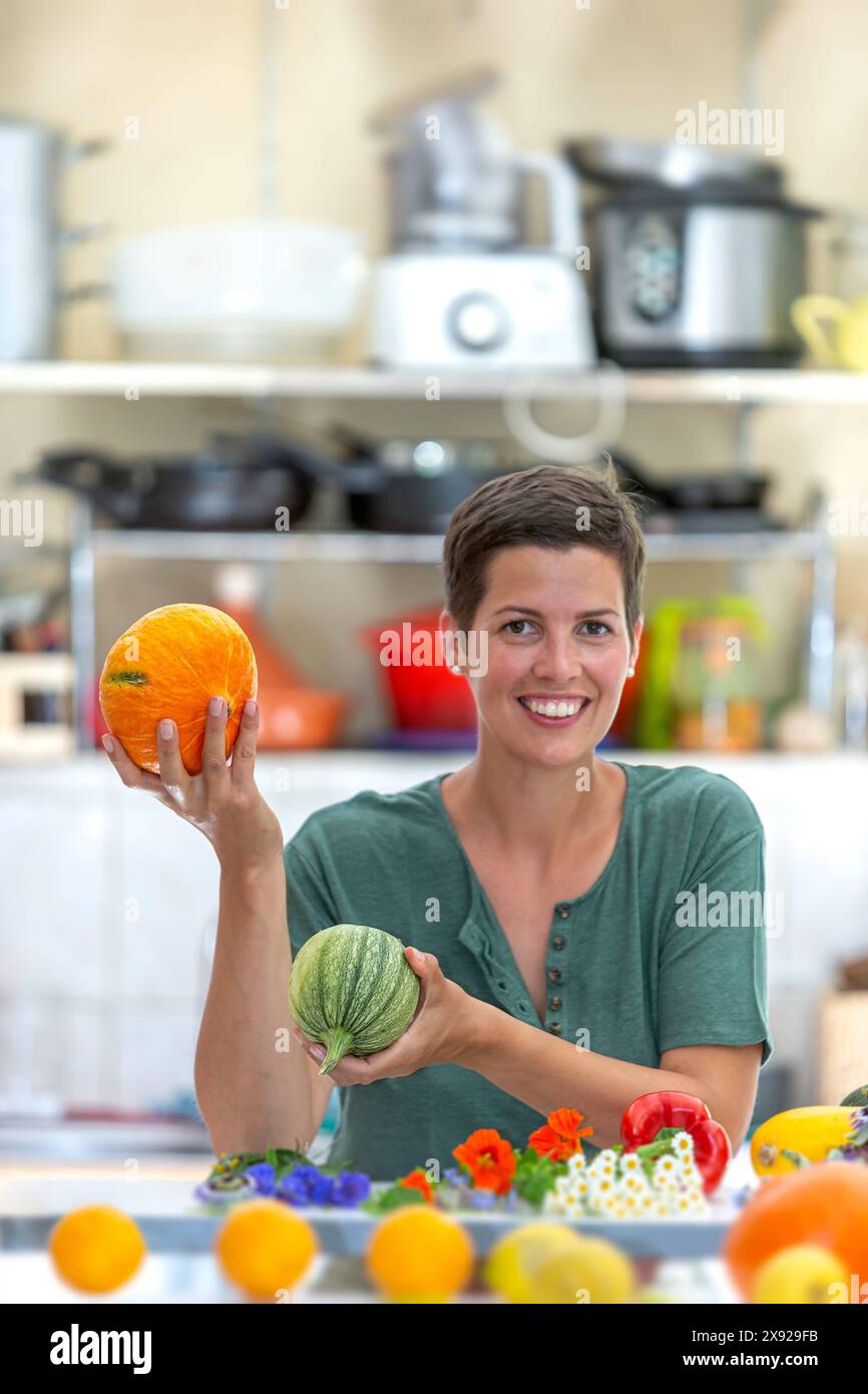 Donna sorridente e rilassata, con un mucchio di verdure biologiche davanti e con zucchine e zucca in mano. Ritratto di una giovane donna nella sua cucina dietro il piano di lavoro. 016643 096 Foto Stock