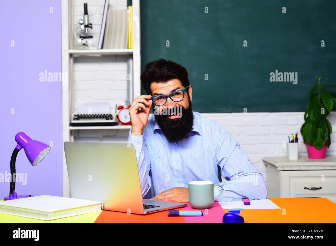 Insegnante di sesso maschile sorridente in classe seduto alla scrivania lavorando su un computer portatile. Studente universitario in occhiali che studia online. E-learning al college o. Foto Stock