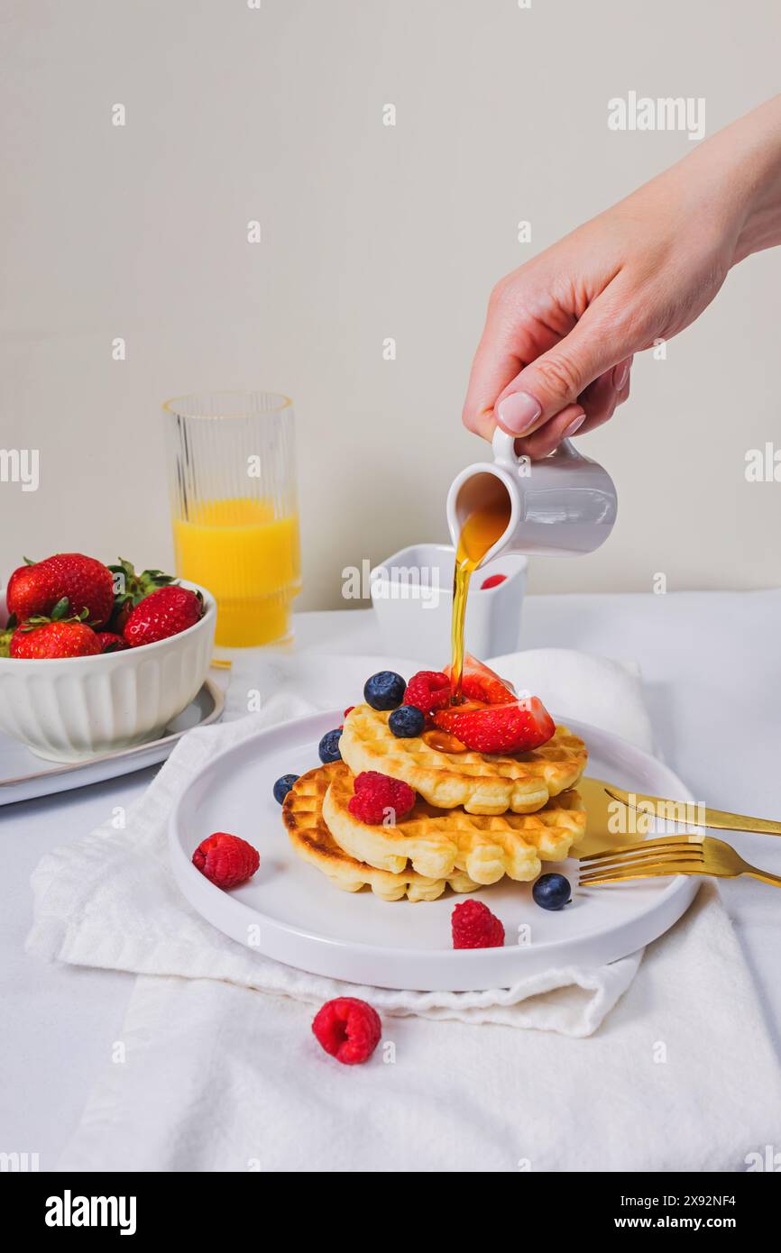 Primo piano della mano femminile che versa sciroppo d'acero dal piattino sopra i dolci waffle fatti in casa con frutti di bosco Foto Stock