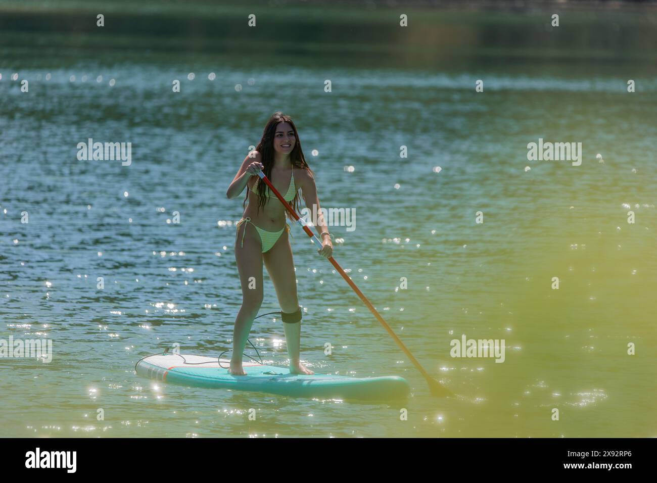 Giovane donna latina in bikini che si gode di paddle surf su un lago, con cespugli che oscurano il primo piano della foto, creando una scena naturale serena Foto Stock