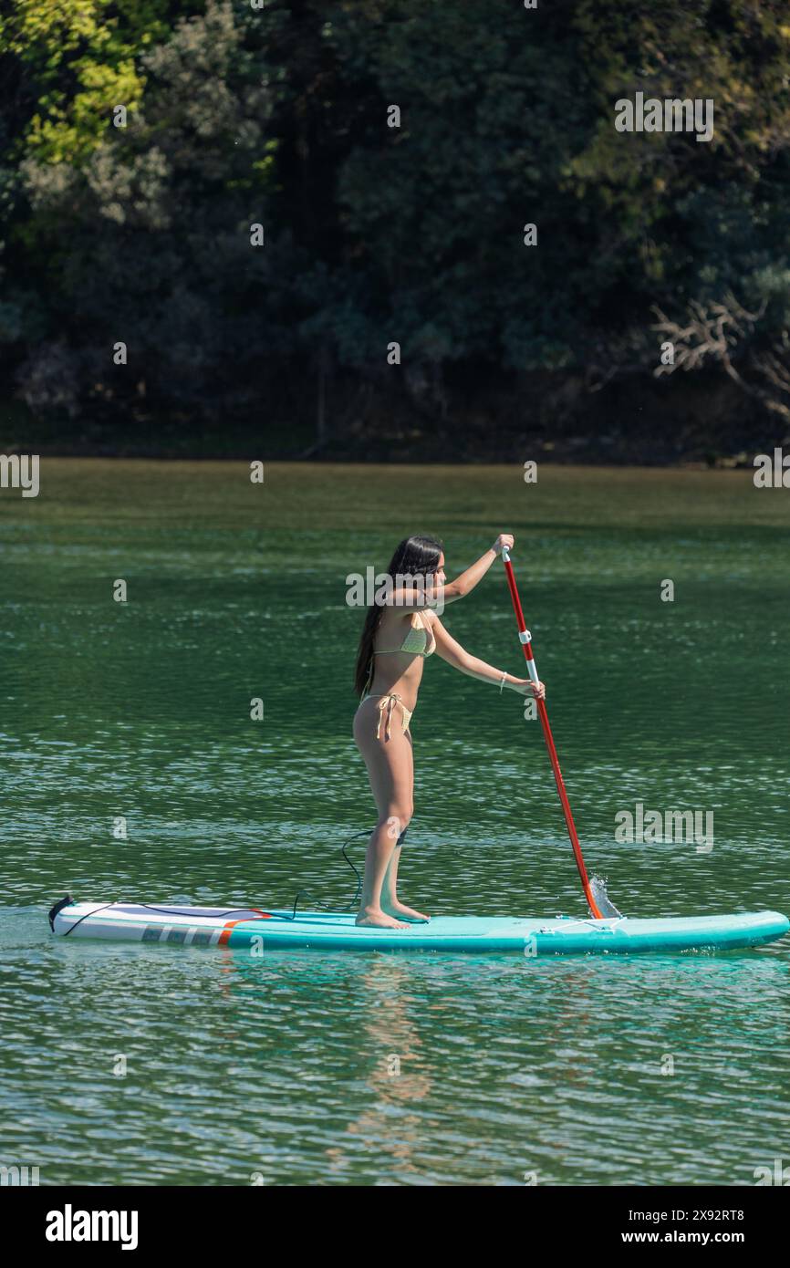 una donna multiculturale verticale si distingue per il paddleboard su un fiume, circondato da una natura lussureggiante. Il suo spirito attivo per gli sport all'aperto a. Foto Stock