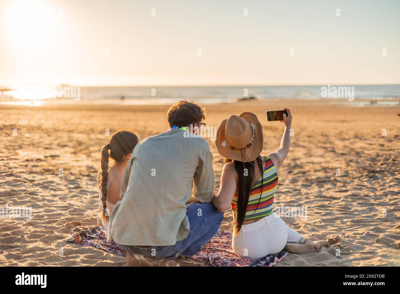 Tre amici multiculturali vengono visti da dietro mentre fanno un selfie con un cellulare sulla spiaggia, guardando il tramonto sul mare. Il cappuccio dell'immagine Foto Stock