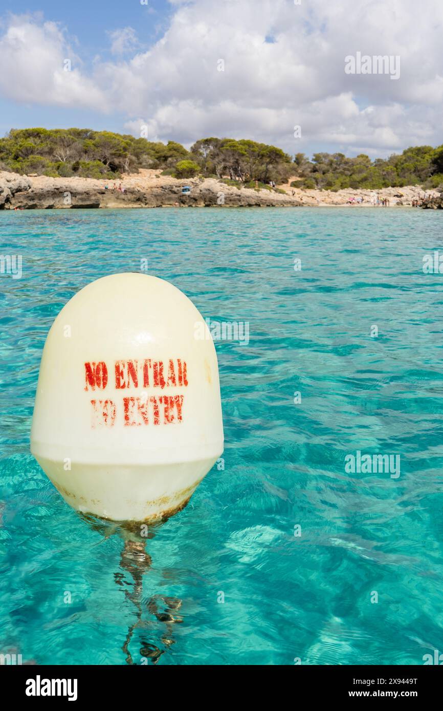 Primo piano di una boa galleggiante nell'acqua del mare con l'avviso "NESSUN INGRESSO" e sullo sfondo una piccola e incontaminata insenatura nell'isola di Minorca, in Spagna. Foto Stock