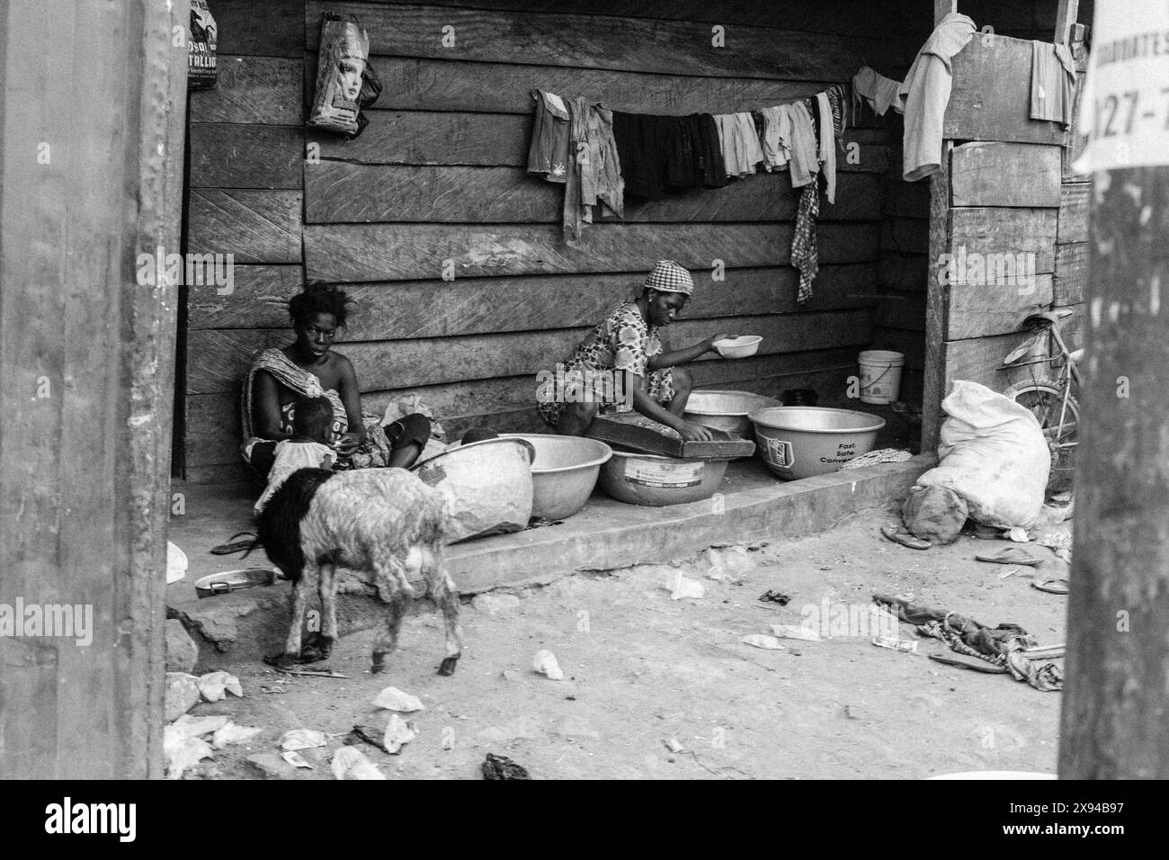 Donne e bambini in Ghana seduti e che lavorano all'esterno di una struttura in legno, con una capra nelle vicinanze, che mette in risalto la vita rurale quotidiana. Foto Stock