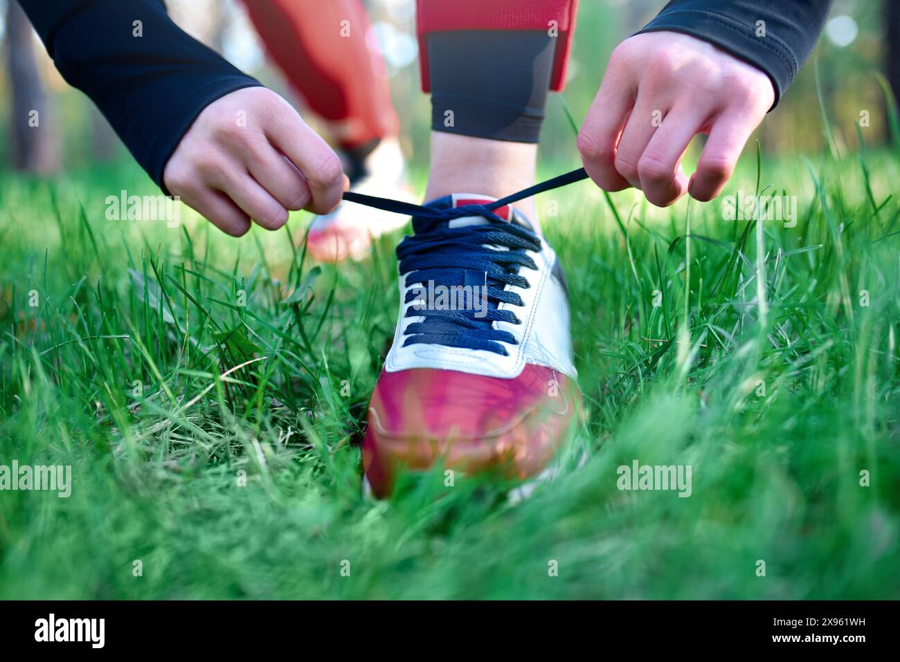 Persona che lega lacci scarpe a una sneaker rossa e bianca, preparandosi per un'attività all'aperto in un'area erbosa. Foto Stock
