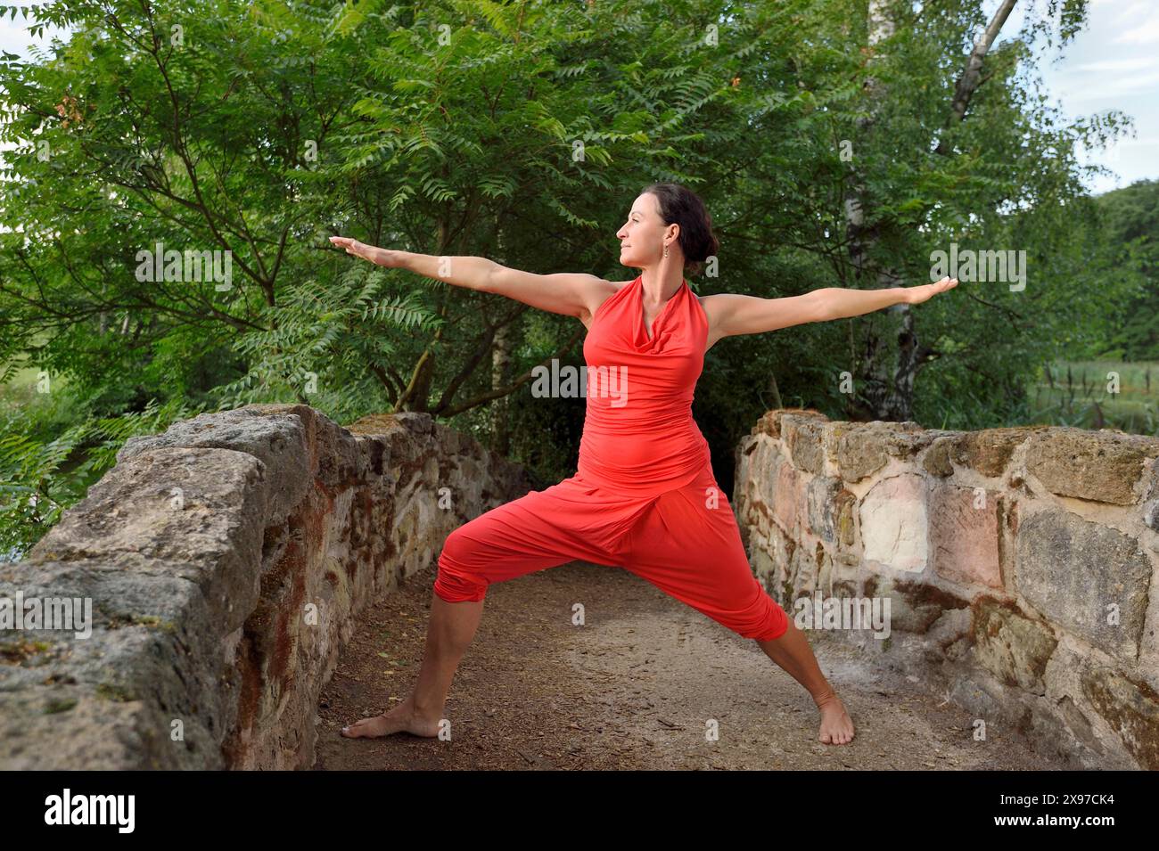 Donna di mezza età in un abito rosso che fa yoga, The Warrior 2 pose, Virabhadrasana II, in un parco in estate, Germania Foto Stock