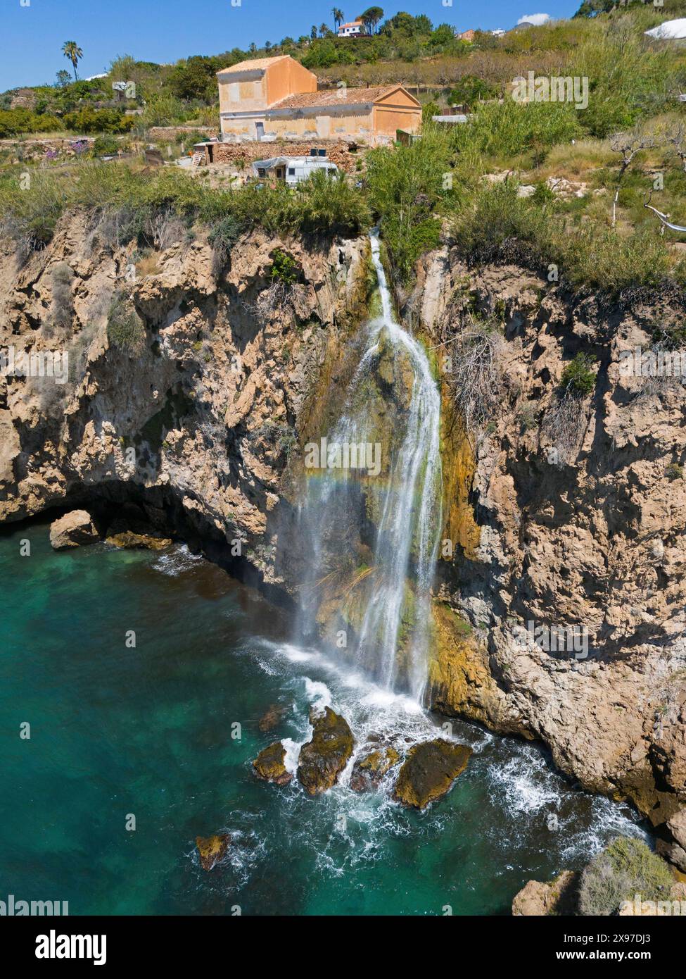 Una pittoresca cascata cade da una scogliera in acque turchesi con case sullo sfondo, un ambiente roccioso naturale, vista aerea, Cascada Foto Stock