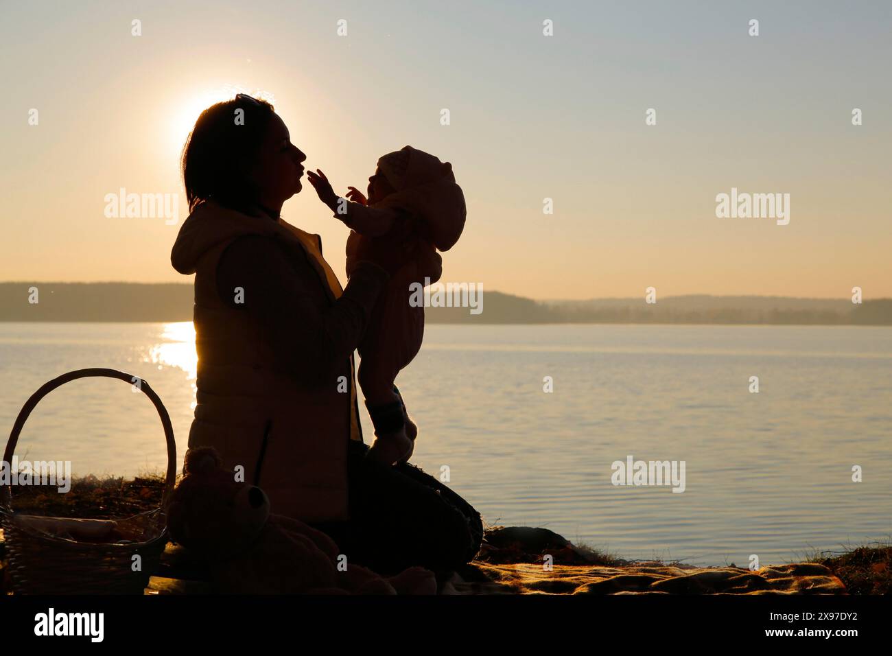 Silhouette di una madre che solleva il bambino vicino a un lago al tramonto, creando un momento tranquillo e sereno di legame Foto Stock