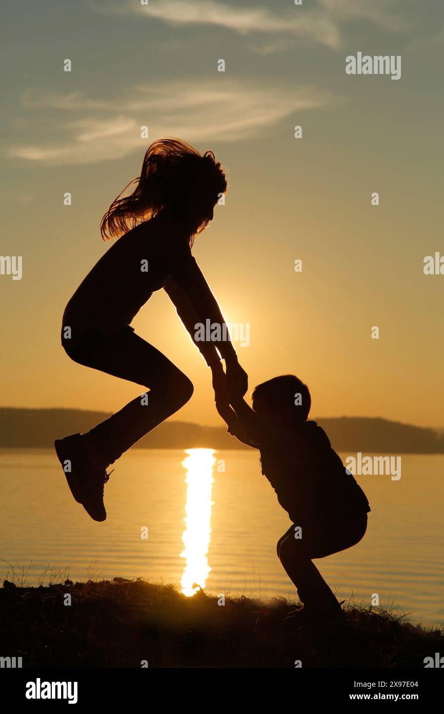 Silhouette di bambini che giocano accanto a un lago al tramonto, creando un'atmosfera serena e giocosa Foto Stock