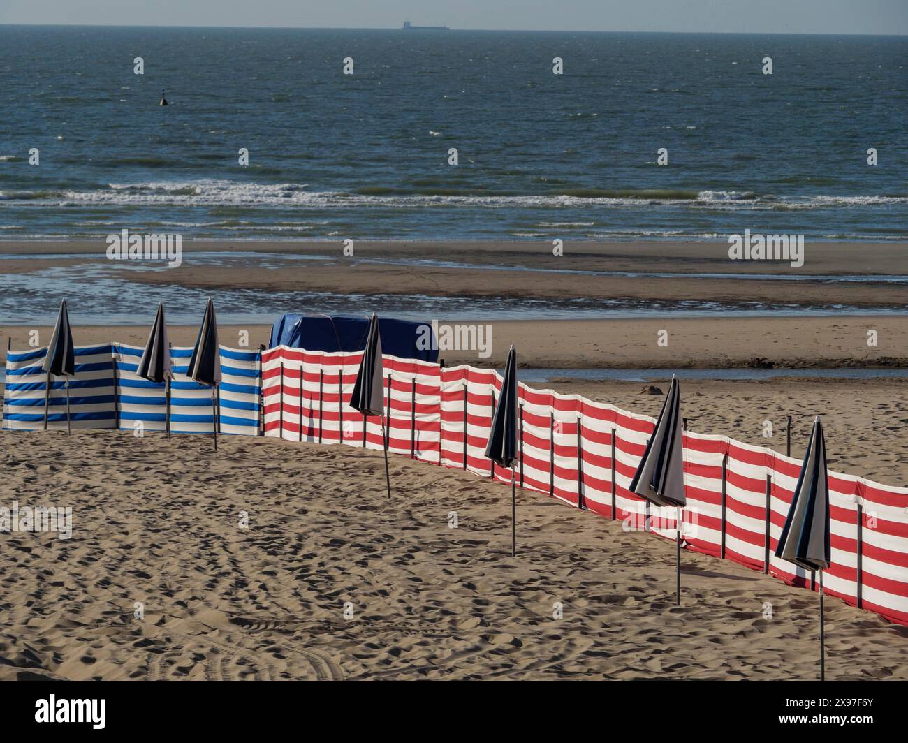 Ampia spiaggia sabbiosa con ombrelloni neri e a strisce blu e bianche sparsi e il mare con onde sullo sfondo, colorate sedie a sdraio Foto Stock