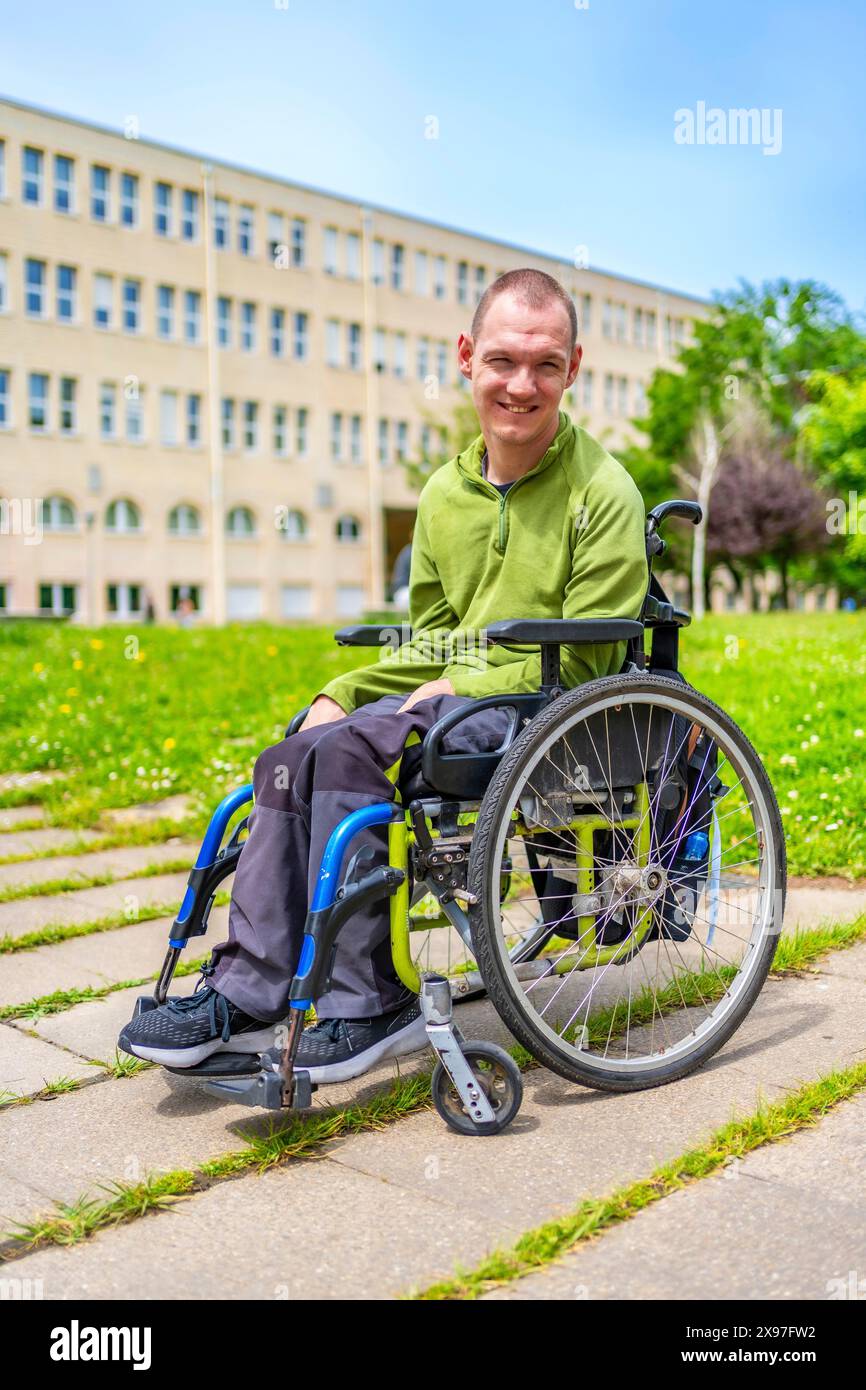 Uomo in sedia a rotelle sorridente alla macchina fotografica fuori dall'università nel campus con erba verde Foto Stock