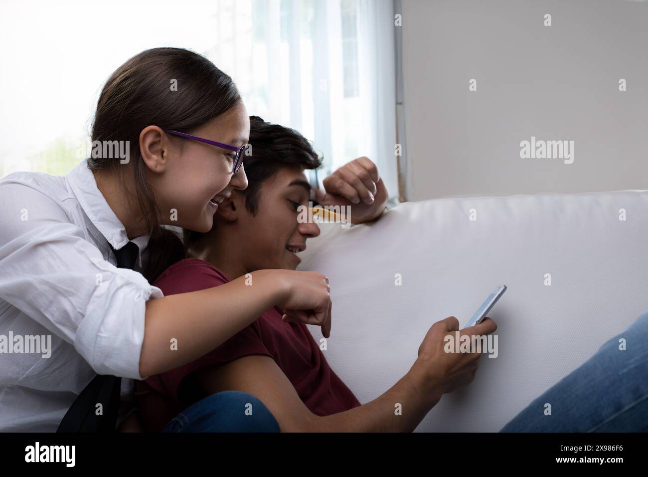 giovane donna con gli occhiali, sorridente calorosamente, si appoggia sulla spalla di un giovane uomo mentre tiene in mano uno smartphone. Sono entrambi seduti su un divano bianco, chiaramente Foto Stock