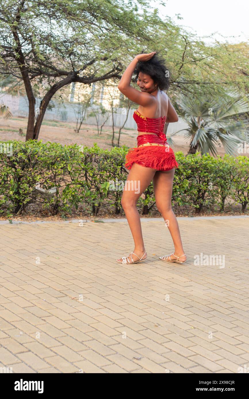 Donna sorridente che balla in costume rosso all'aperto, vista posteriore, capelli ricci, movimento espressivo. Foto Stock