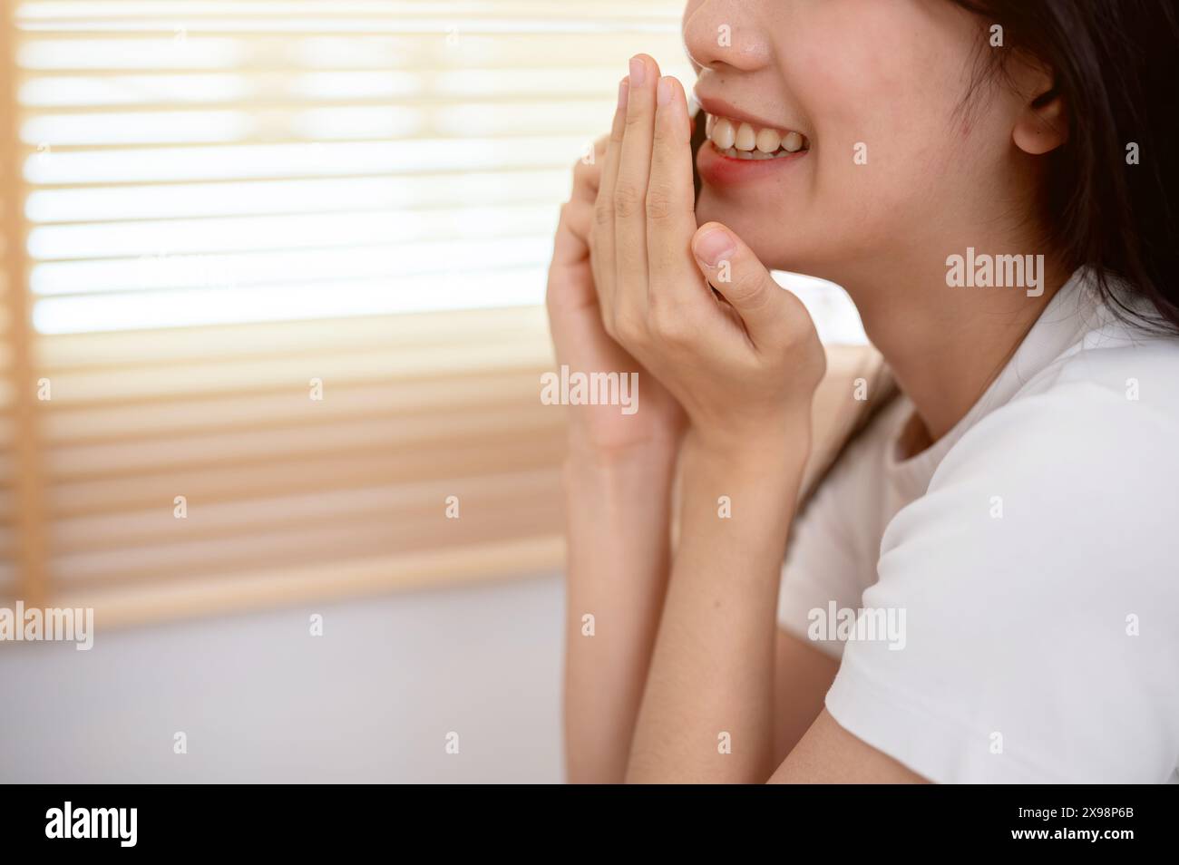 Immagine ritagliata di una giovane donna asiatica allegra che si copre la bocca mentre parla al telefono con la sua amica in casa, si diverte a parlare o a chiacchierare Foto Stock