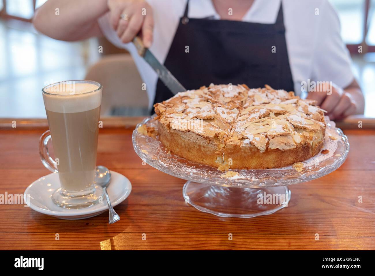 Panettiere che taglia una torta rotonda in spugna posizionata su un banco per torte di vetro, atmosfera accogliente e una donna professionale che serve una deliziosa torta Foto Stock