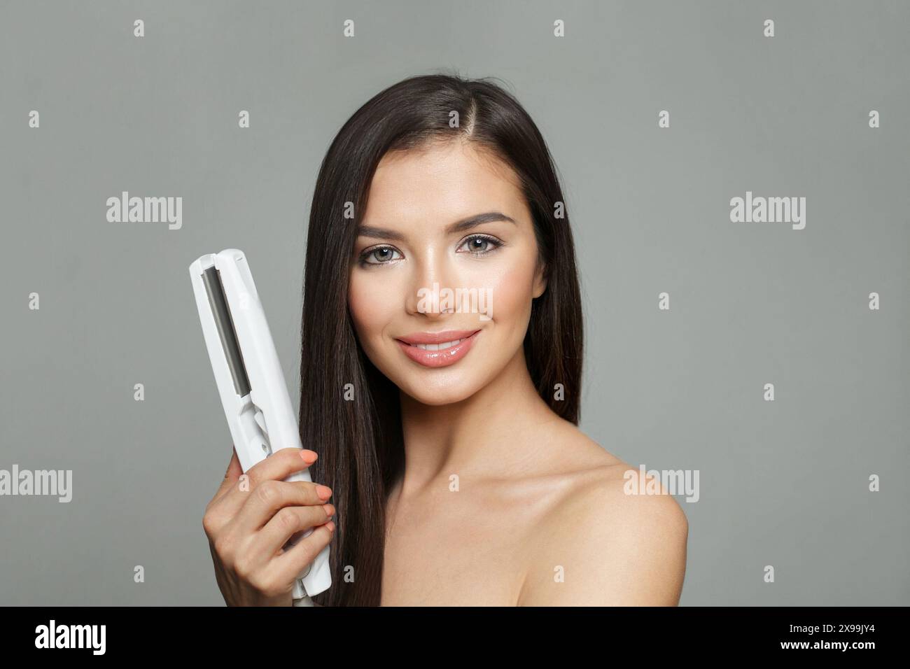 Concetto di lisciatura dei capelli. Giovane donna bruna in salute che tiene il ferro da stiro per capelli e lisciando i suoi lunghi capelli luminosi e lisci su sfondo bianco Foto Stock