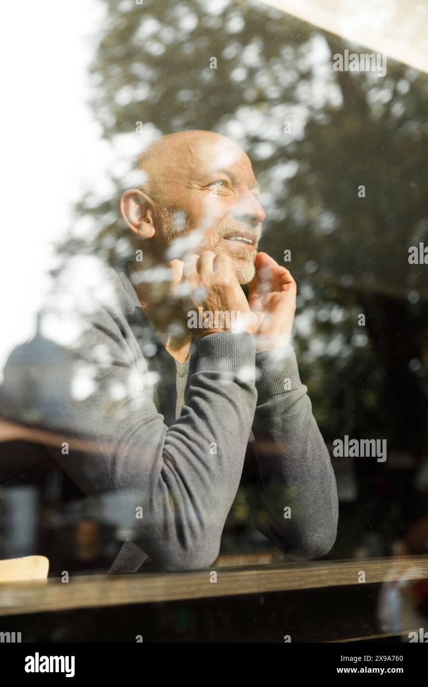 Allegro anziano con la barba che guarda in alto e sorride attraverso una finestra con riflessi Foto Stock