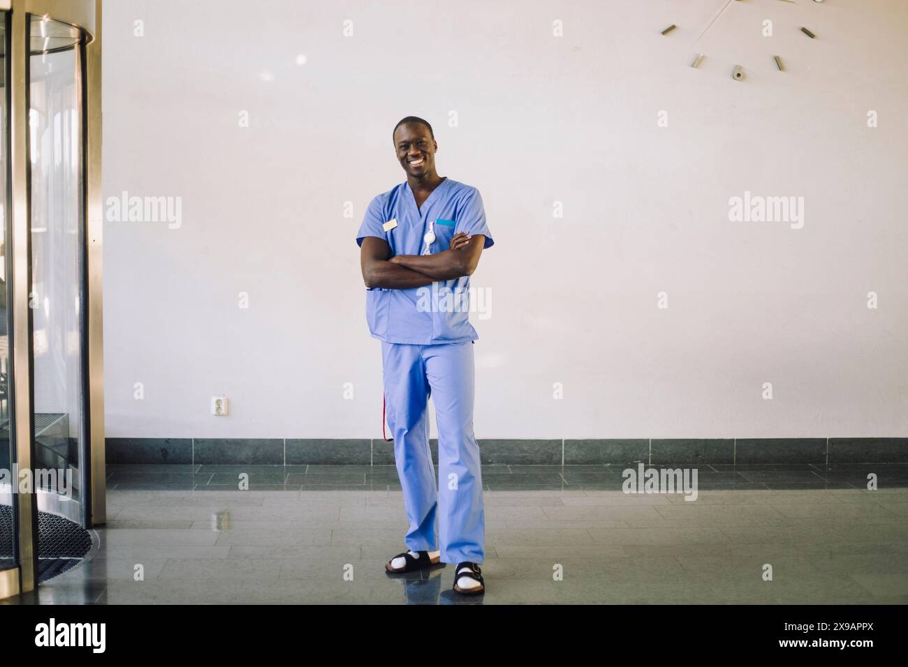 Ritratto di medico maschio sorridente in piedi con le braccia incrociate di fronte a parete bianca all'ospedale Foto Stock