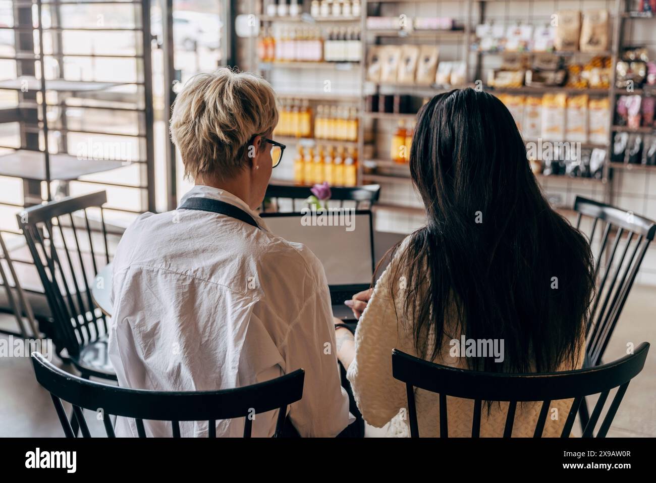 Vista posteriore della proprietaria e della collega seduta al tavolo che lavora con un computer portatile nel bar Foto Stock