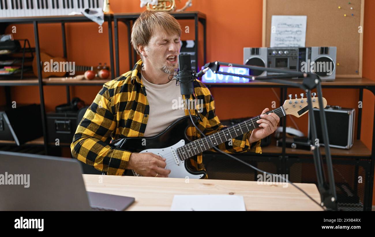 Un giovane uomo biondo e barbuto suona la chitarra elettrica e canta in un microfono in uno studio di musica, proiettando emozioni e musiche. Foto Stock