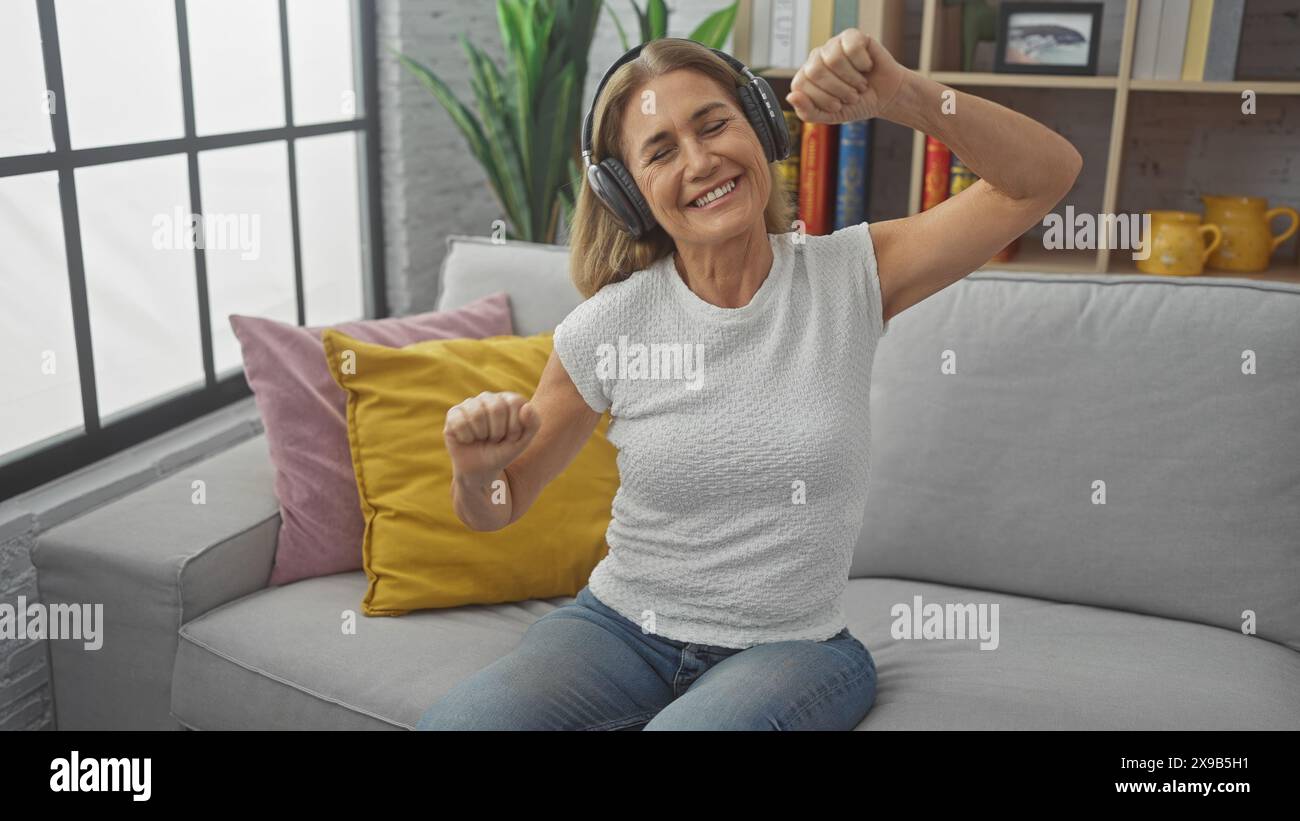 Una donna gioiosa di mezza età che indossa le cuffie balla su un divano in un salotto accogliente ascoltando musica. Foto Stock